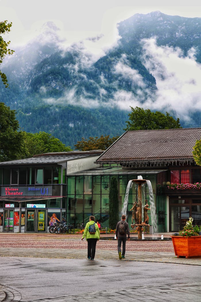 Rainy day in a town with a fountain, theater, and misty mountain backdrop.